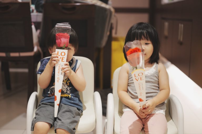 弟弟與姊姊各拿一支玫瑰花擋在臉前，坐在小椅子上